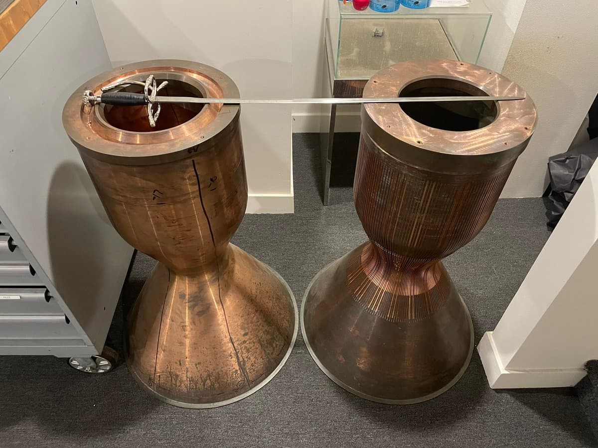 Two large, copper-colored Tesla coils stand side-by-side on a gray carpeted floor, with a long metal rod connecting their top terminals. The coils feature a distinctive hourglass shape with vertical ridges and are positioned in an indoor setting with white walls and a glass display case in the background.