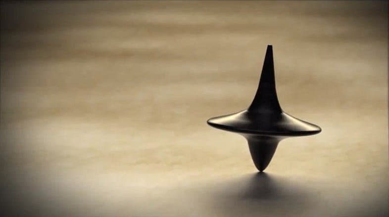 A black, metallic, abstract sculpture resembling a stylized spinning top or a droplet of liquid metal stands on a smooth, light-colored surface. The object features a wide, flat central disc with a tall, tapering spike rising from the center and a smaller, inverted cone at the base.