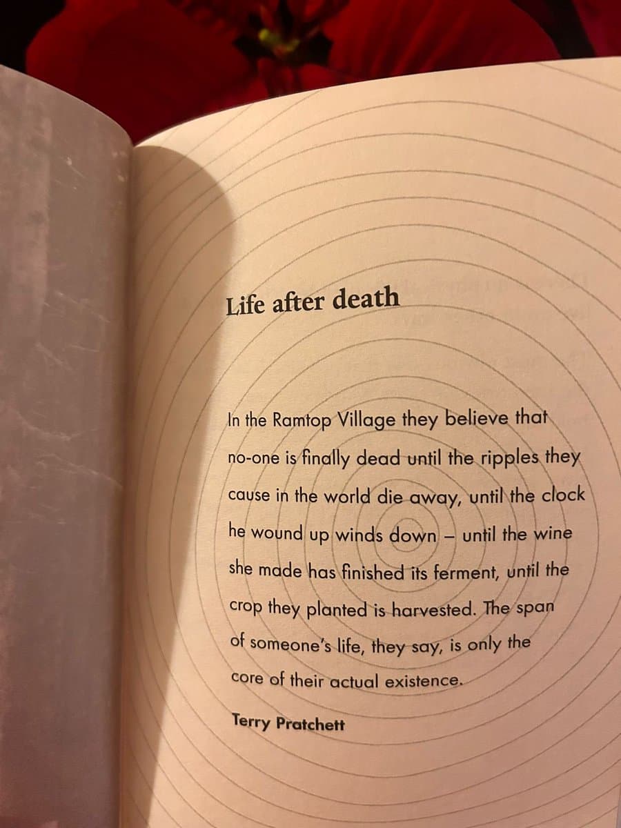 A photograph of an open book page featuring a quote about life after death, set against a background of red flowers. The page has a design of concentric circles radiating from the center.