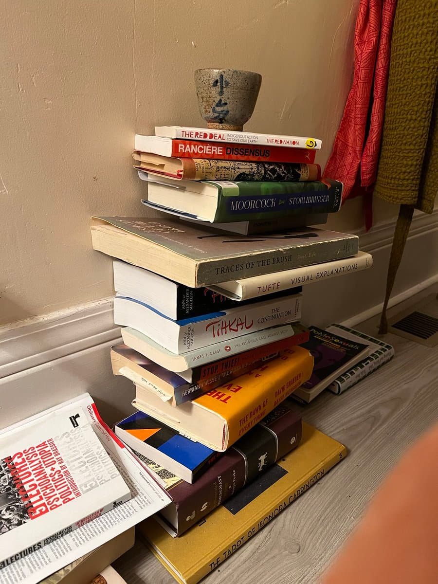 A photograph showing a tall, precarious stack of books on a wooden floor, with a ceramic cup balanced on top. The scene is set against a beige wall with a red and green curtain visible in the background.