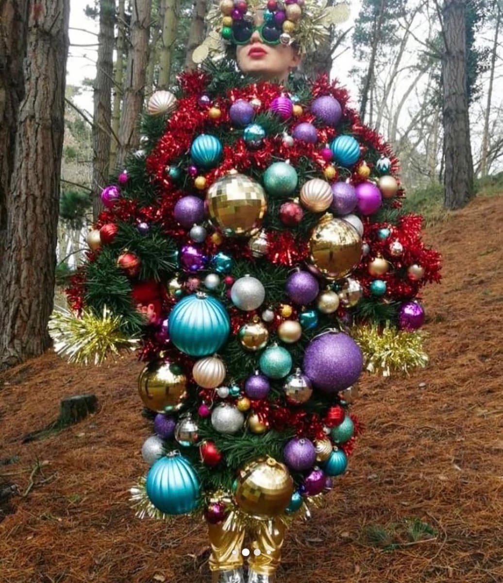A person stands in a forest wearing an elaborate costume made of green pine branches, red tinsel, and numerous colorful Christmas ornaments, including large gold, purple, and blue baubles. They are also wearing gold pants and a headpiece with ornaments and sunglasses.