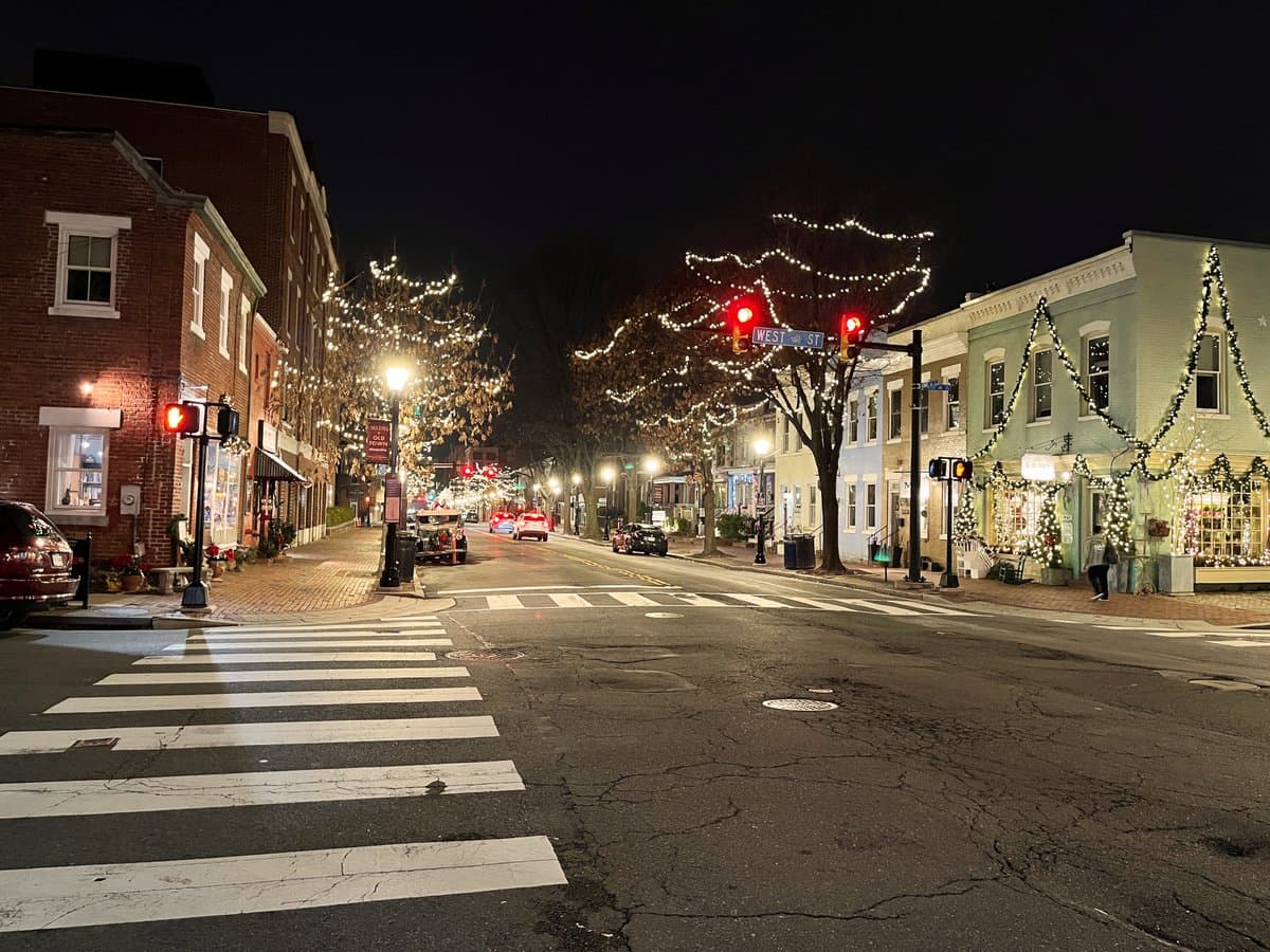A nighttime street scene in a small town, featuring brick and painted buildings decorated with festive holiday lights and garlands. The street is lined with illuminated trees and traffic lights, with a crosswalk in the foreground and a few cars visible in the distance.