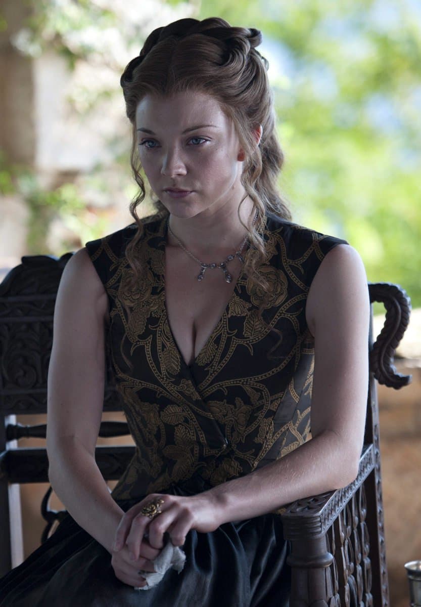 A woman with reddish-brown hair styled in an updo sits on an ornate wooden chair, wearing a black and gold patterned dress and a necklace. She holds a small white cloth in her hands and looks off to the side with a serious expression. The background is a blurred outdoor setting with greenery.
