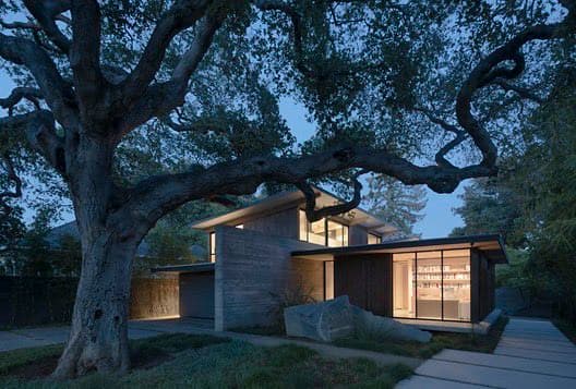 A modern, single-story house with large glass windows and a concrete exterior is illuminated at dusk, nestled beneath the sprawling branches of a large, mature tree. The scene features a paved walkway leading to the entrance and a landscaped yard with a prominent boulder.