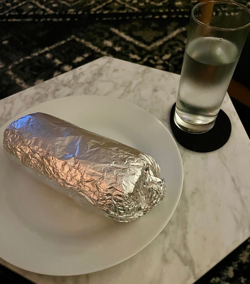 A photograph of a meal consisting of a foil-wrapped item on a white plate and a glass of water on a black coaster, set on a marble table. The background features a dark, patterned rug.