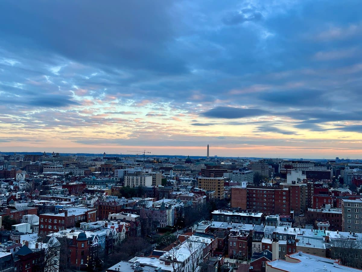 A panoramic cityscape photograph taken from an elevated vantage point, showing a dense urban area with brick buildings and snow-dusted rooftops under a dramatic, cloudy sky at dusk or dawn. A tall, thin obelisk is visible on the distant horizon.