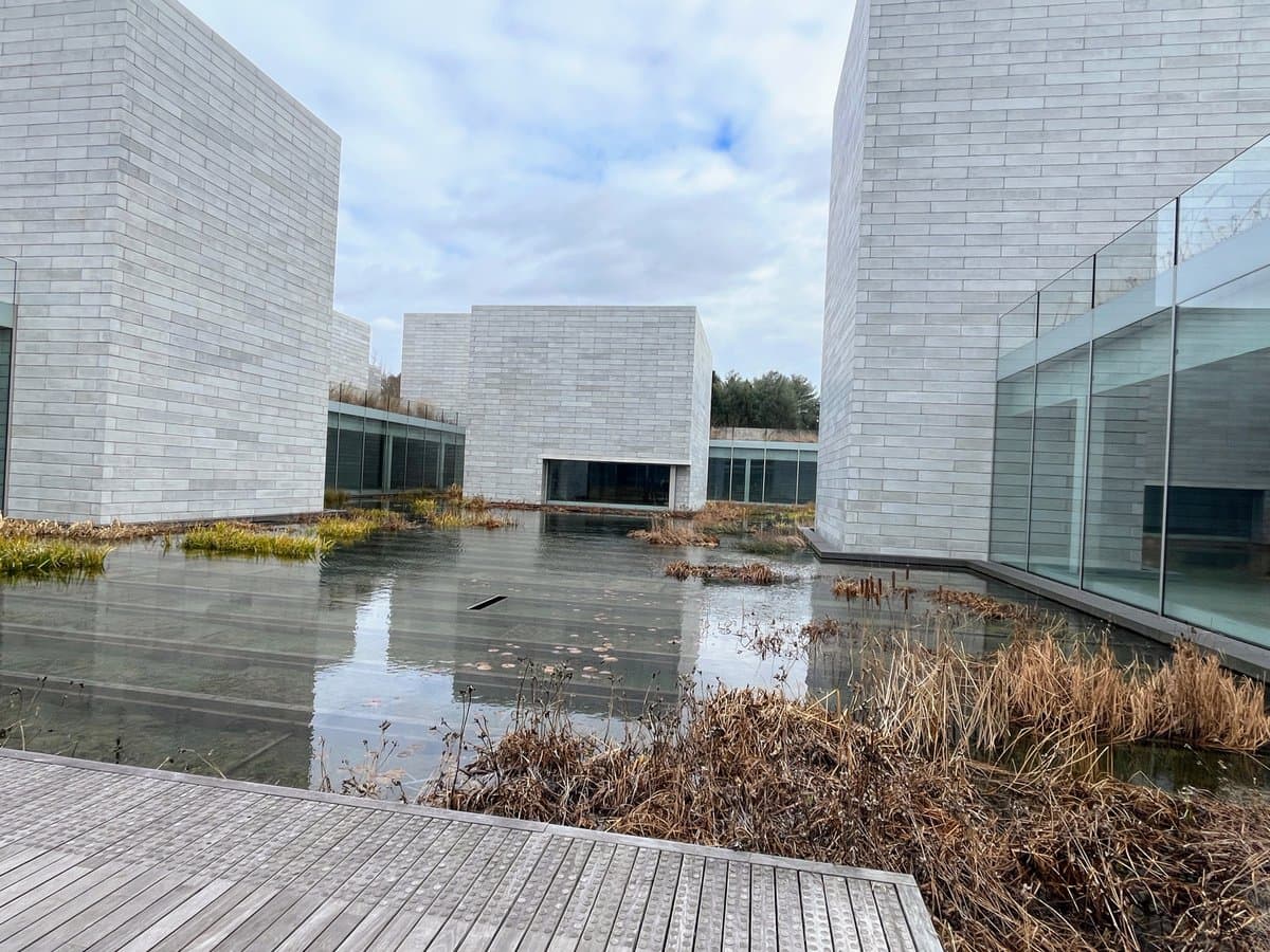 A photograph of a modern architectural complex featuring white stone buildings surrounding a calm, reflective pond with dried reeds and a wooden walkway in the foreground.