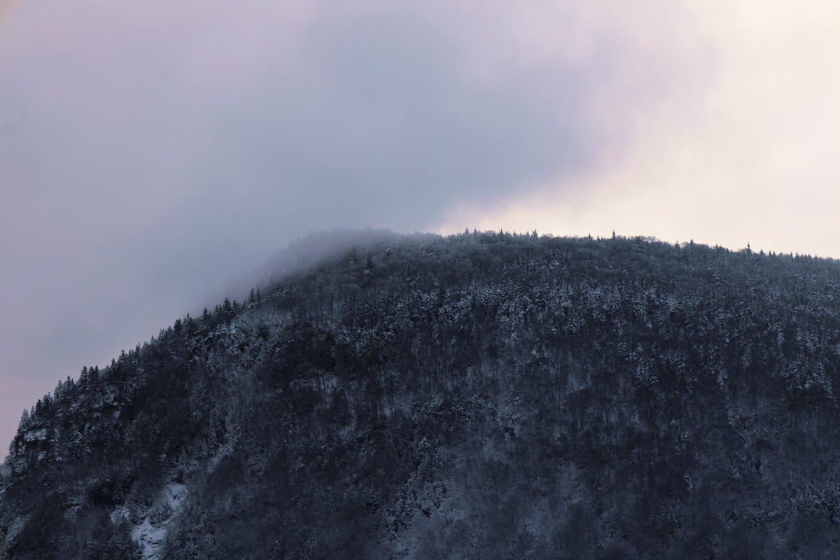 A landscape photograph showing a snow-covered, forested mountain ridge partially obscured by low-hanging clouds and fog under a pale, overcast sky.