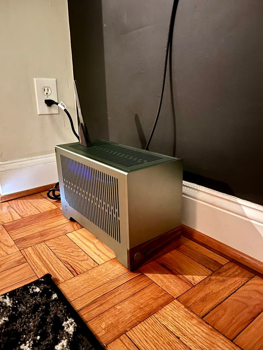 A photograph of a silver Mac Pro computer with a wooden base sitting on a parquet floor next to a wall outlet.