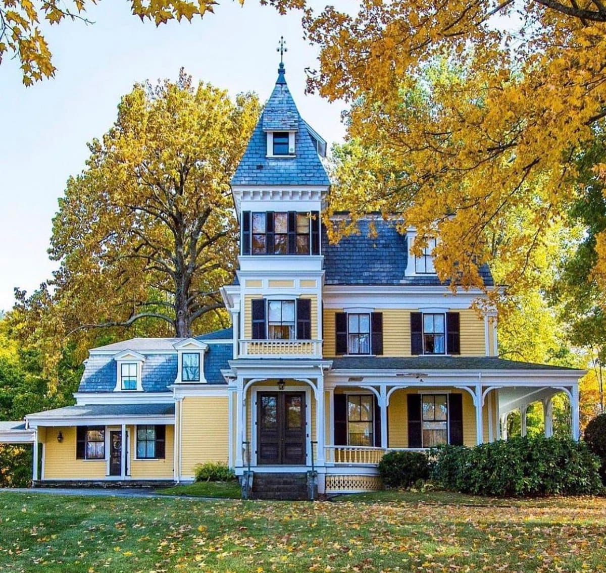 A yellow, multi-story Second Empire style house with a prominent blue-slate roof and a central tower, set in a grassy yard surrounded by trees with autumn foliage.