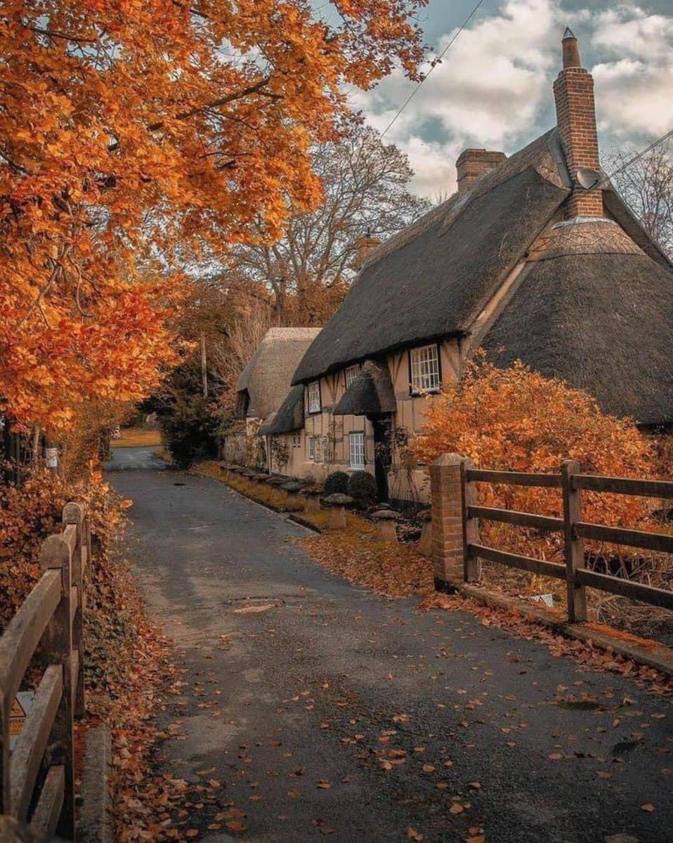A scenic photograph of a traditional English village in autumn, featuring a thatched-roof cottage with a brick chimney, a narrow paved road, and trees with vibrant orange foliage.