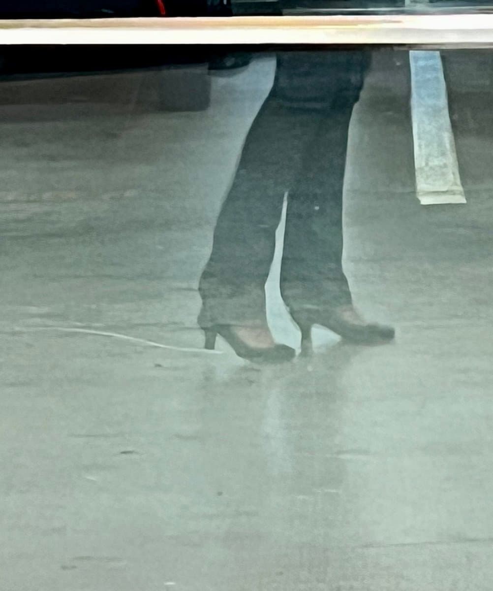A low-angle, grainy photograph showing a person's lower legs and feet wearing dark pants and high-heeled shoes while standing on a concrete surface.
