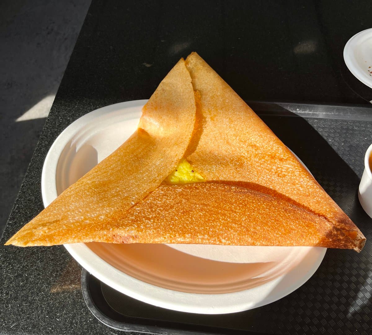 A folded, golden-brown dosa with a visible yellow filling sits on a white disposable plate, which is placed on a black tray.