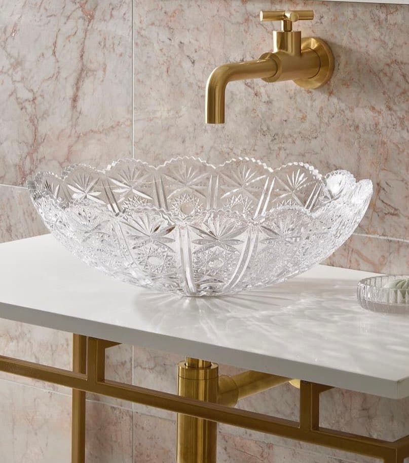 A crystal glass vessel sink with an intricate cut-glass pattern sits on a white marble countertop, paired with a gold wall-mounted faucet and a gold-framed vanity. The background features a light-colored marble wall.