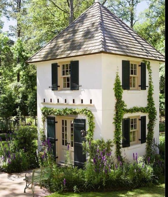 A small, two-story white house with a shingled roof and dark green shutters, surrounded by lush greenery and purple flowers. The structure features a front door with sidelights and is adorned with climbing vines on its exterior walls.