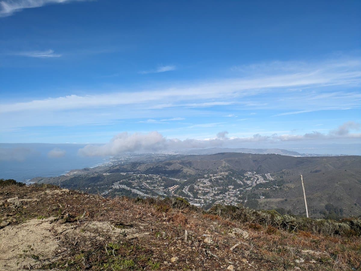 A scenic landscape photograph taken from a high vantage point, looking down over a coastal valley with hills, a town, and the ocean in the distance under a blue sky with scattered clouds. The foreground shows dry, rocky terrain with sparse vegetation.