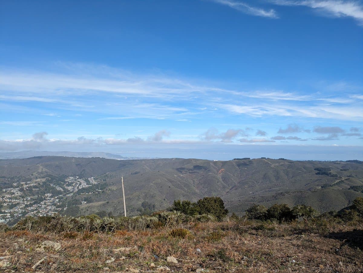 A wide landscape photograph taken from a hilltop, showing rolling green and brown hills under a bright blue sky with wispy clouds. In the distance, a hazy coastline and a body of water are visible, with a residential area nestled in the valley to the left.