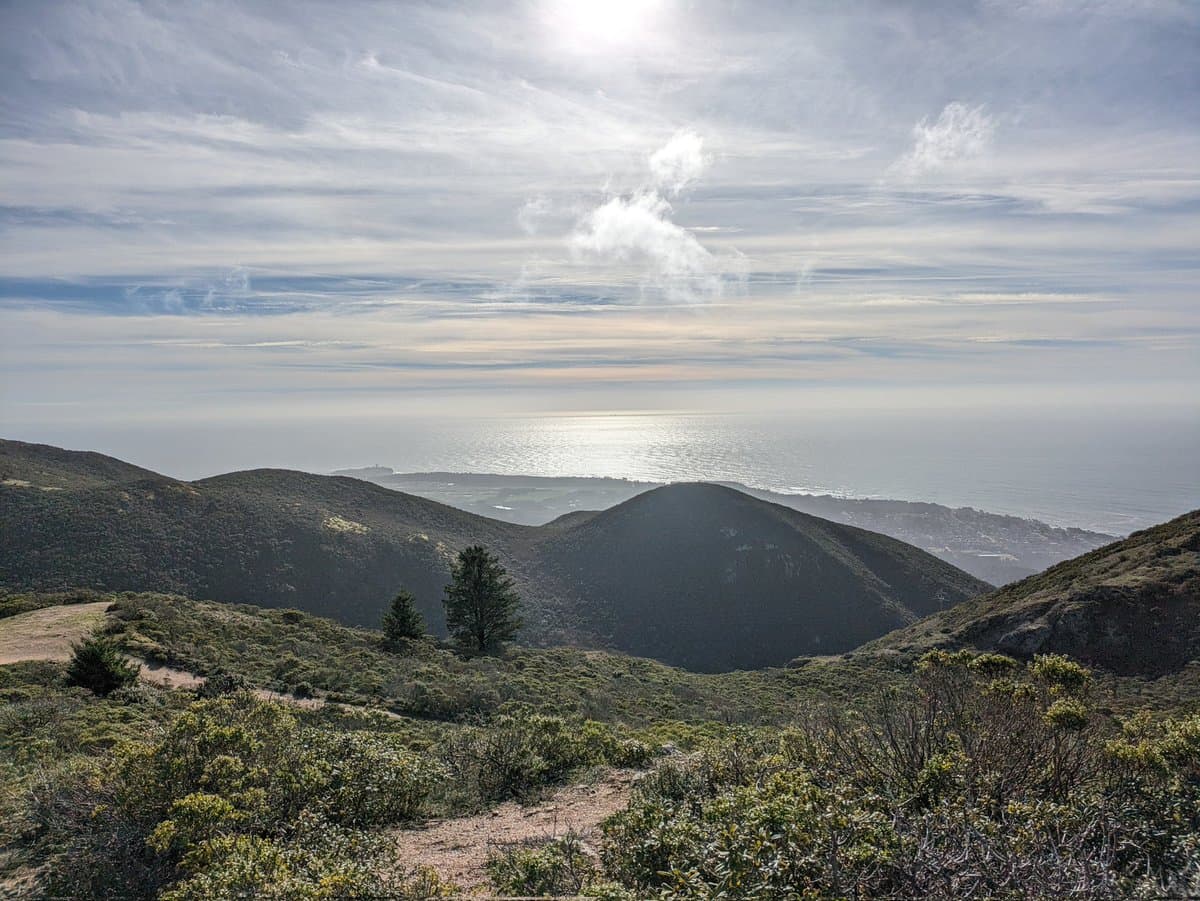 A scenic landscape photograph showing a view from a hilltop overlooking a coastal region. The foreground features a dirt trail winding through low-lying shrubs and grasses on a hillside, with rolling hills in the midground leading to a coastline and the ocean in the distance under a partly cloudy sky.
