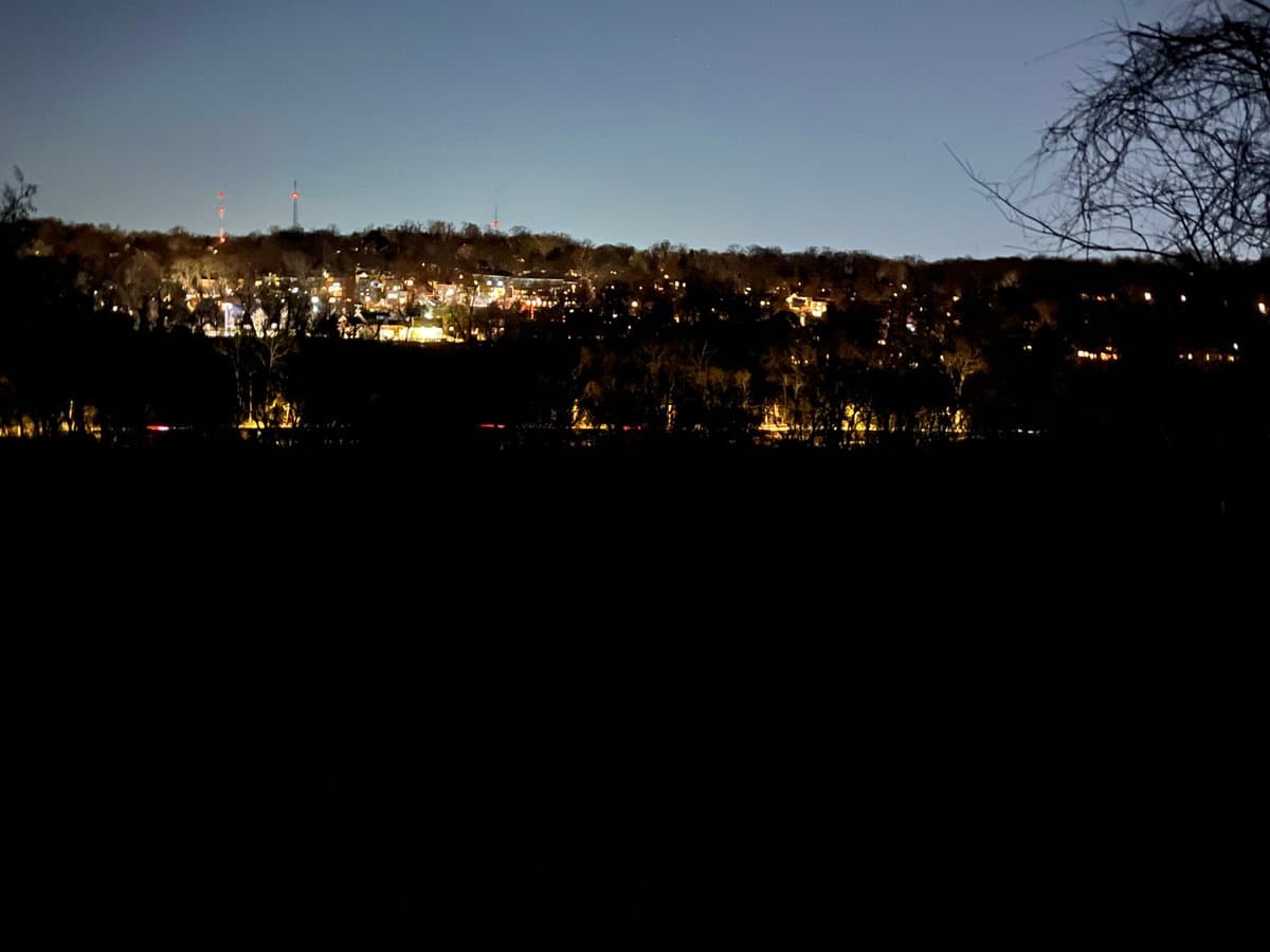 A nighttime landscape photograph showing a dark foreground with a distant, illuminated hillside and city lights under a twilight sky. Bare tree branches are visible on the right side of the frame.