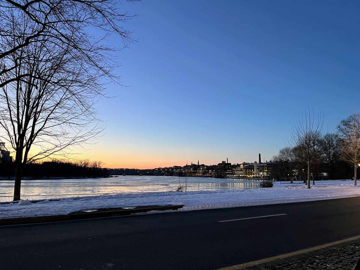 A winter landscape photograph showing a snow-covered riverbank and road in the foreground, with a city skyline and bridge visible across the water under a twilight sky. Bare trees frame the scene on the left and right sides.