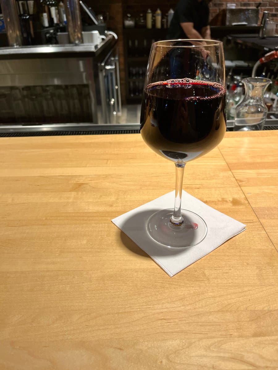 A glass of red wine sits on a white napkin on a wooden bar counter, with a blurred bar background featuring bottles and equipment.