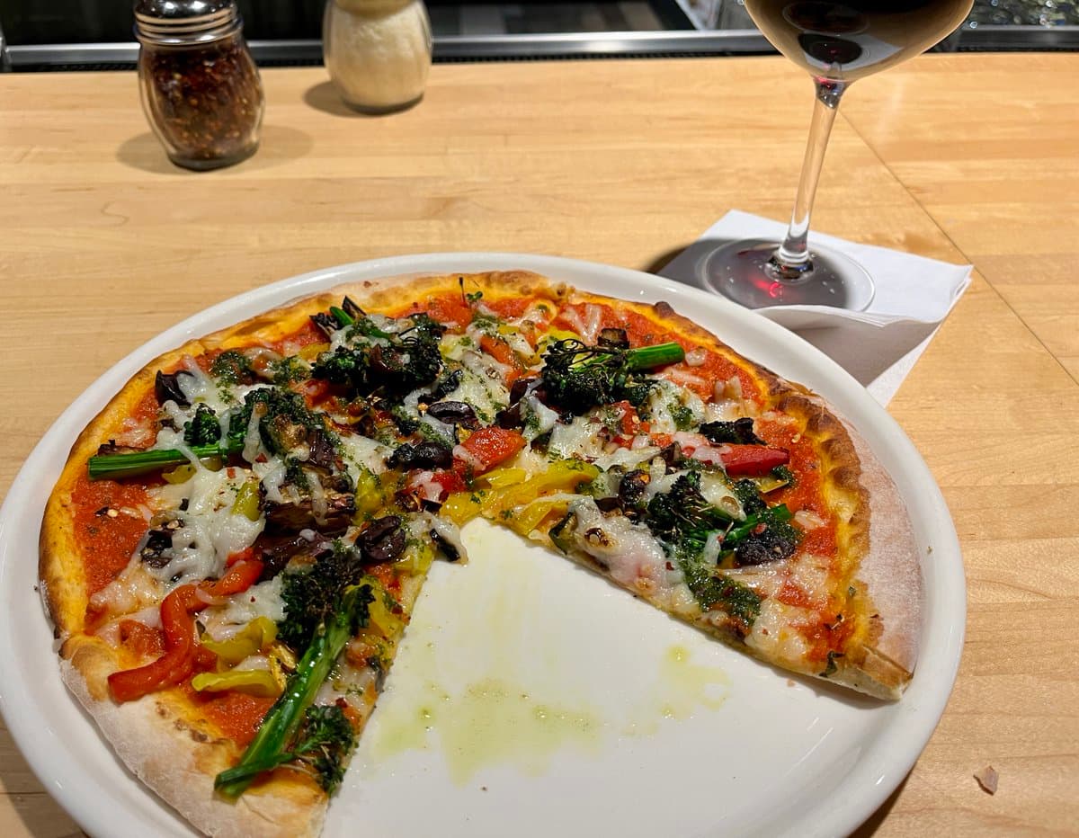 A partially eaten pizza topped with broccoli, peppers, and cheese sits on a white plate on a wooden table, accompanied by a glass of red wine and condiment shakers.