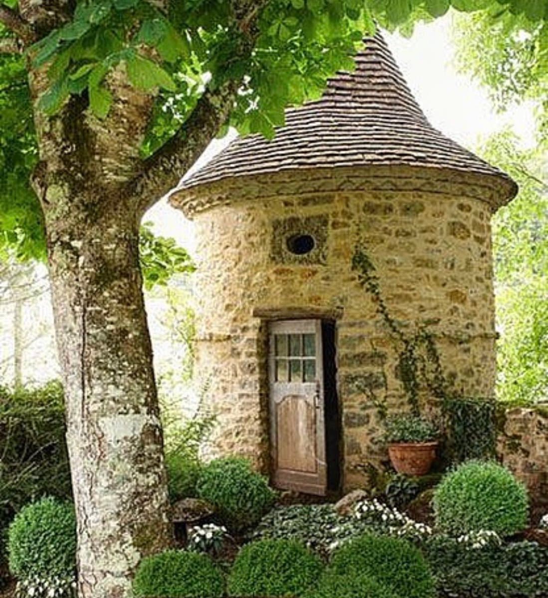 A small, round stone structure with a conical roof, resembling a miniature house or dovecote, sits in a garden setting. It features a small wooden door and a circular opening near the top, surrounded by greenery and a large tree.