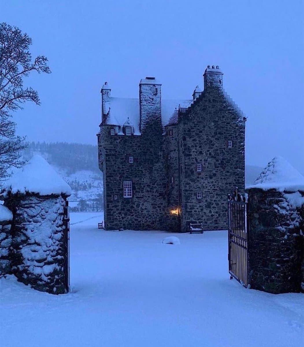 A photograph of a large, historic stone castle covered in snow during a winter storm. The scene is framed by snow-capped stone gate pillars and a metal gate in the foreground, with a single warm light glowing from a window in the building.