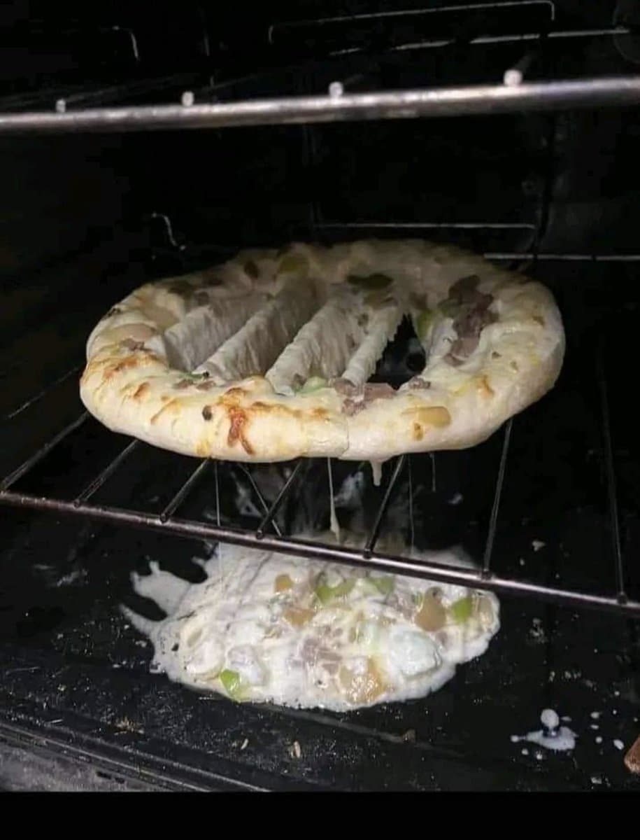 A photograph of a pizza inside an oven, where the center of the pizza has collapsed and fallen through the rack, leaving only the crust and a few strips of dough hanging down. The melted cheese and toppings have pooled on the bottom of the oven below the rack.
