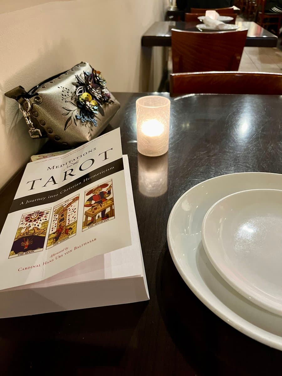 A photograph of a book titled "Meditations on the Tarot" resting on a dark wooden table next to a lit candle, a white plate, and a decorative handbag.