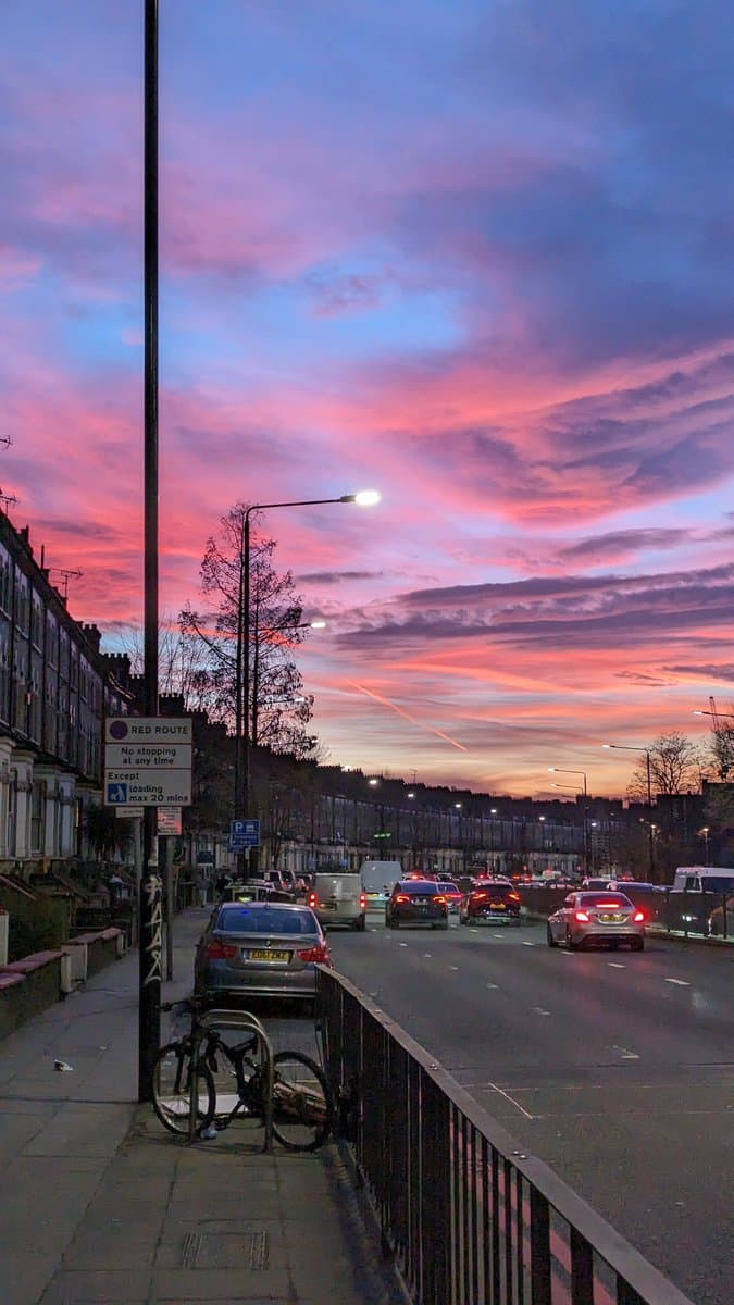 A street scene in London at dusk, featuring a vibrant pink and purple sunset sky over a road with traffic and parked cars. A bicycle is locked to a pole on the sidewalk next to a 'RED ROUTE' traffic sign.