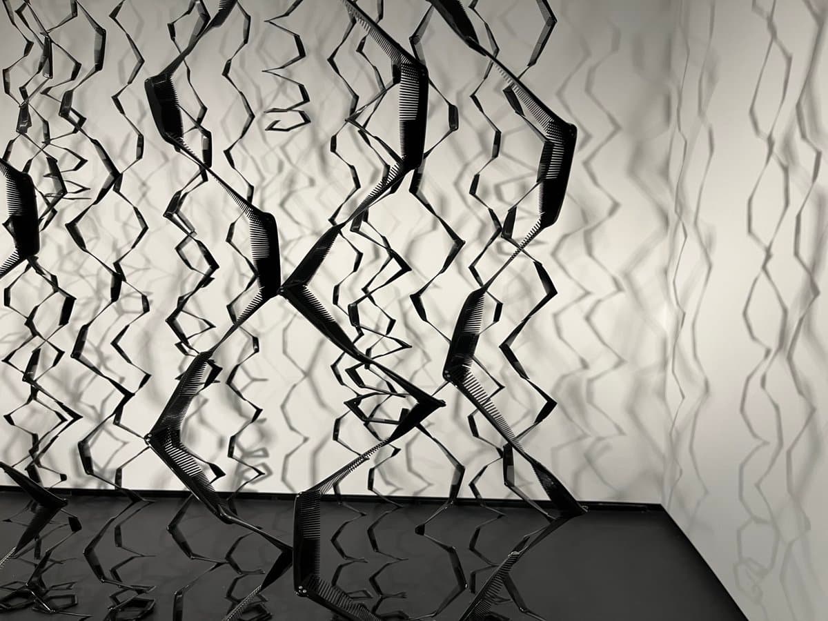 A black-and-white photograph of an abstract art installation featuring a suspended, three-dimensional lattice of dark, zigzagging ribbons. The structure casts intricate, wavy shadows on the white walls and floor of the gallery space.