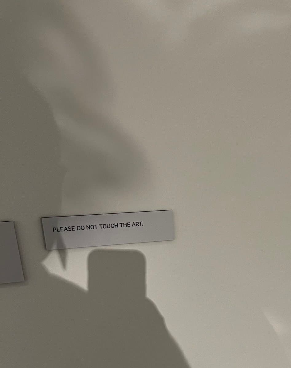 A close-up photograph of a small, rectangular white sign attached to a plain, light-colored wall. The shadow of the person taking the photo is cast on the wall below the sign.