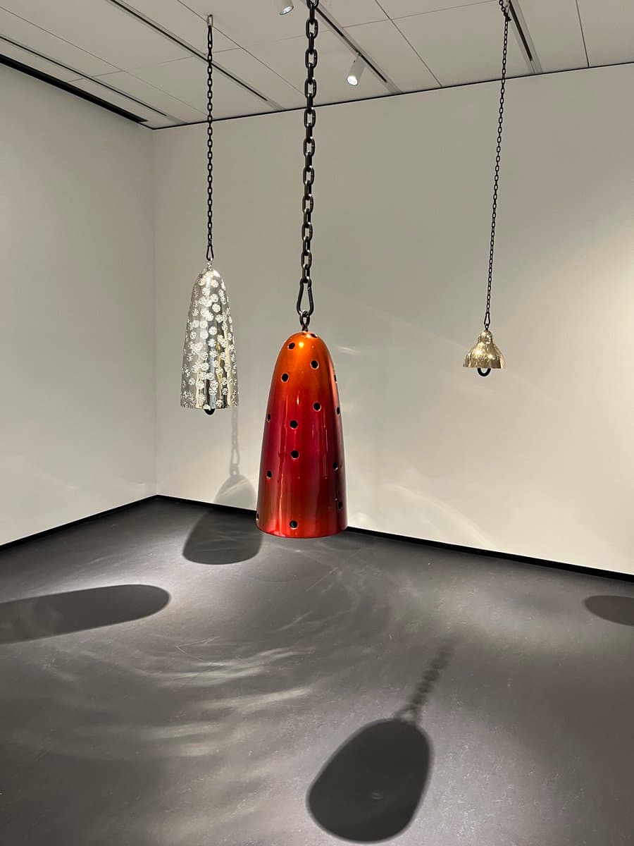 A photograph of a gallery installation featuring three bell-shaped sculptures suspended from the ceiling by black chains. The central object is a large, glossy red-orange bell with perforations, flanked by a silver, textured bell on the left and a smaller, gold-colored bell on the right. Their shadows are cast on the dark floor below.