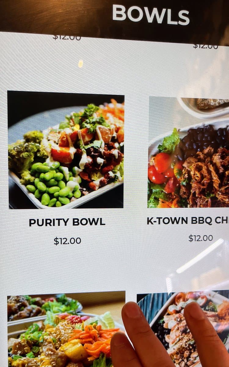 A close-up photograph of a restaurant menu board displaying food items under the heading 'BOWLS'. The image shows a photo of a 'PURITY BOWL' containing broccoli, edamame, and other vegetables, priced at $12.00, next to a 'K-TOWN BBQ CH' bowl. A person's finger is visible at the bottom of the frame.