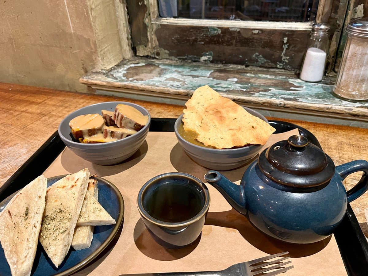 A tray of Middle Eastern food served on a wooden table, featuring bowls of hummus and a soup with flatbread, a plate of pita bread, and a blue ceramic teapot with a cup. The setting includes a rustic window sill with salt and pepper shakers in the background.