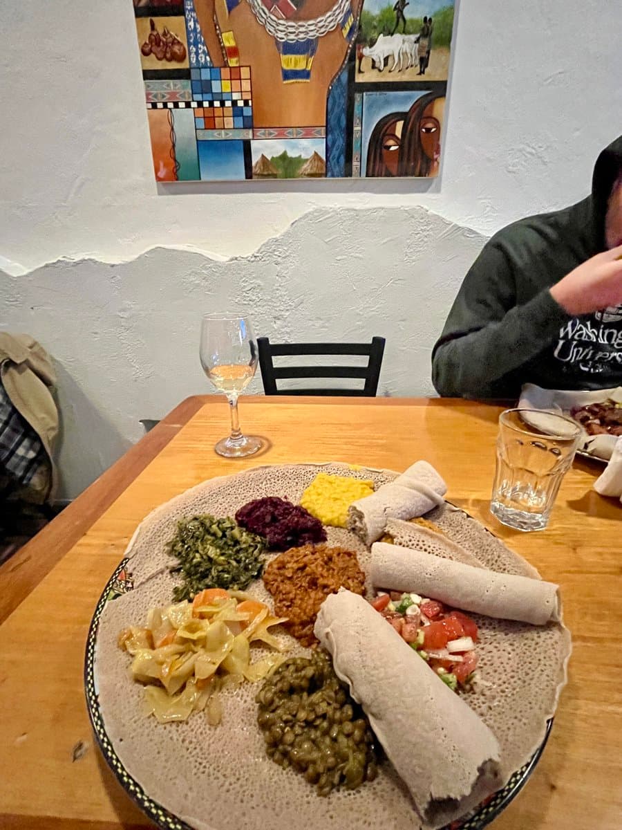 A large platter of Ethiopian food, featuring injera bread with various stews and rolled wraps, sits on a wooden table in a restaurant. A person is seated at the table, and a colorful, mosaic-style painting hangs on the wall behind them.