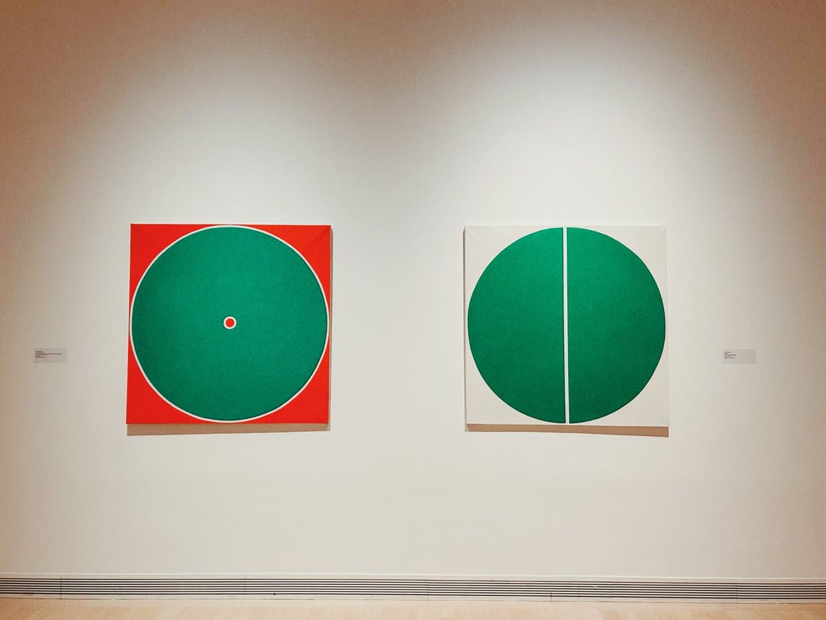 A photograph of two abstract paintings hanging side-by-side on a white gallery wall. The left painting features a large green circle on a red square background, while the right painting shows a green circle split vertically on a white square background.