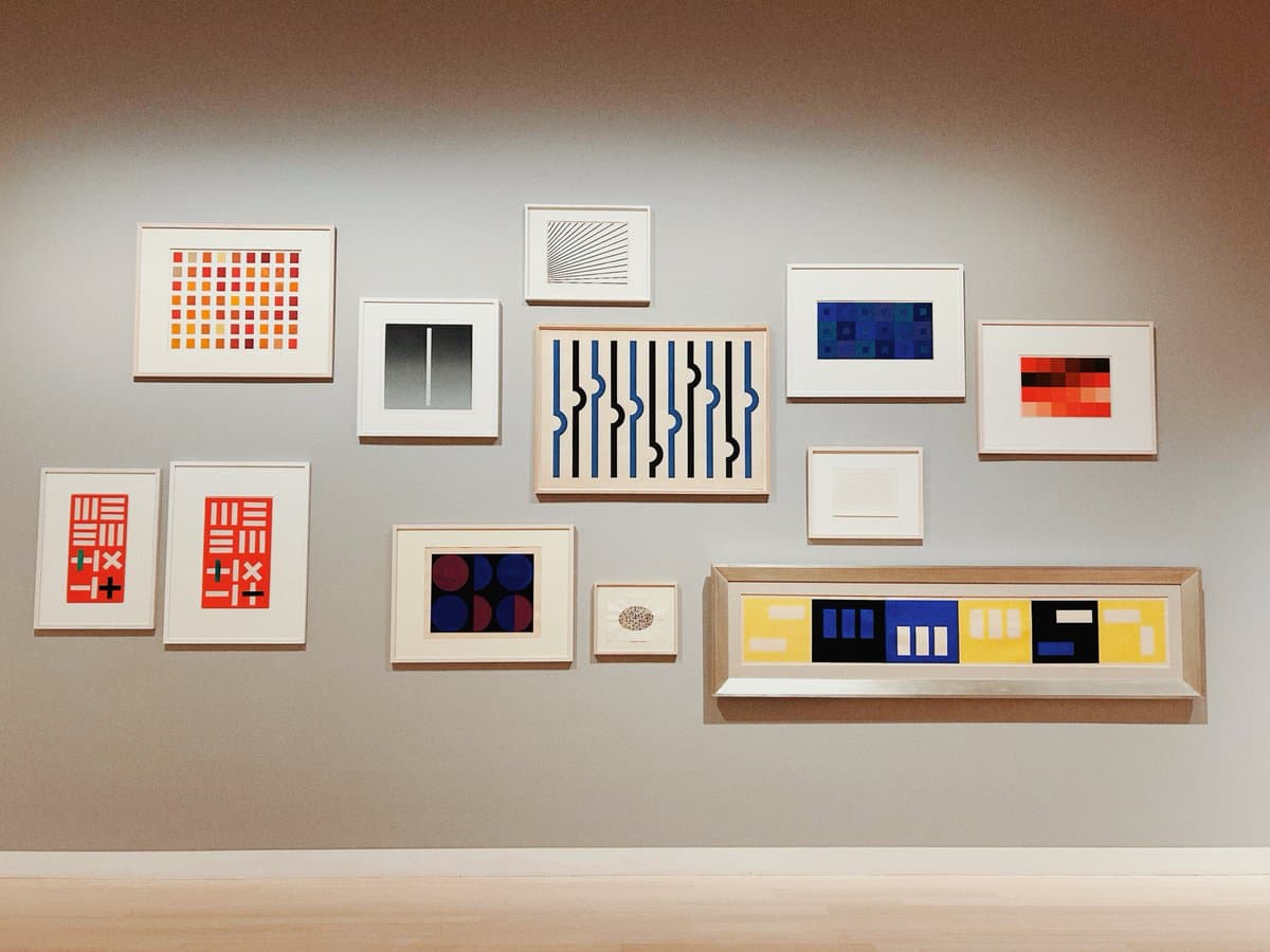 A gallery wall displaying a collection of framed abstract artworks featuring geometric patterns, grids, and bold colors in red, blue, yellow, and black.