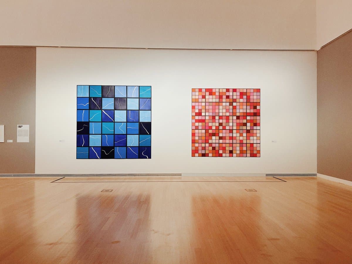 A wide shot of a gallery interior featuring two large, square abstract paintings hanging on a white wall. The painting on the left consists of a grid of blue and black squares with white lines, while the painting on the right is a grid of red, orange, and white squares.