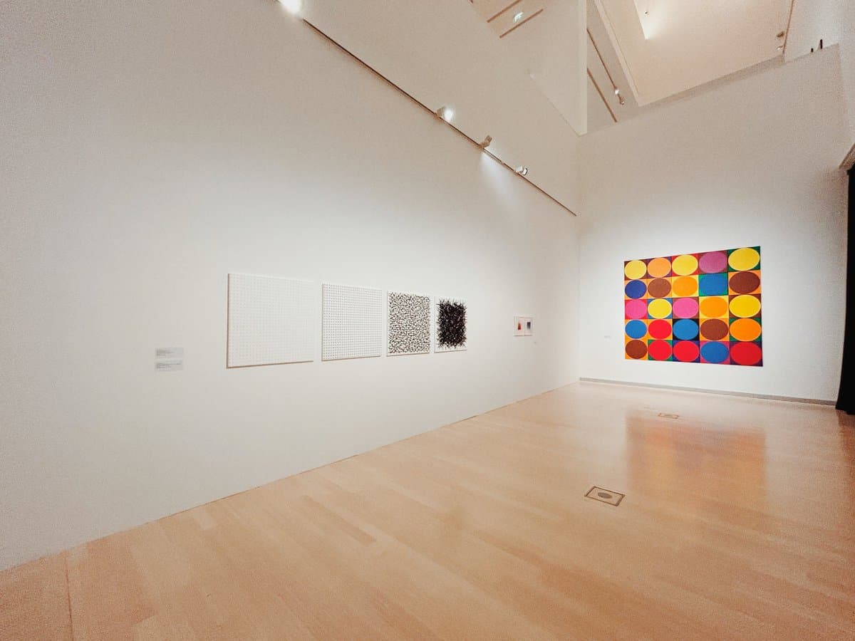 A wide-angle photograph of a bright, empty art gallery with white walls and a light wood floor. Several artworks are displayed, including a series of four square panels on the left wall and a large, colorful grid of circles on the far wall.