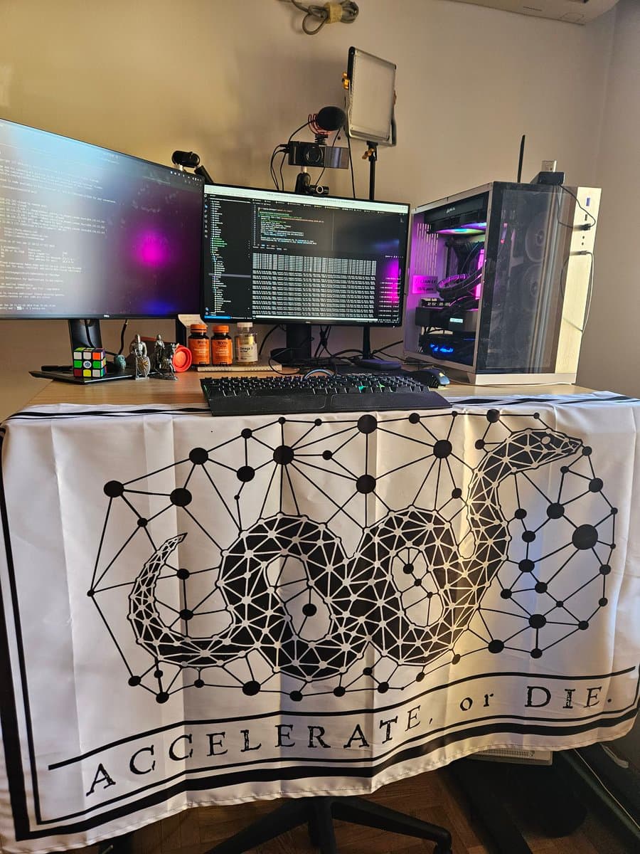 A photograph of a high-performance computer setup featuring a white PC case with internal RGB lighting, two monitors displaying code, and a desk covered by a white banner. The banner features a geometric, low-poly illustration of a snake and the text 'ACCELERATE, or DIE.'