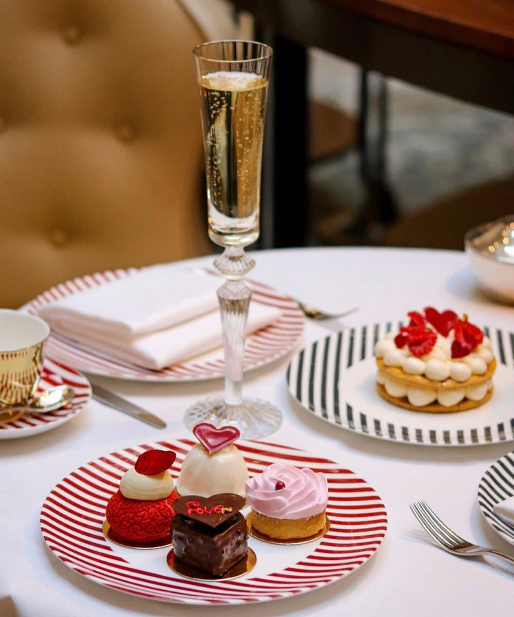 A table setting featuring an assortment of small, heart-themed desserts on a red-striped plate, a glass of sparkling wine, and a larger meringue tart on a black-striped plate. The scene is set with white tablecloths, striped plates, and elegant silverware, suggesting a romantic or celebratory dining experience.