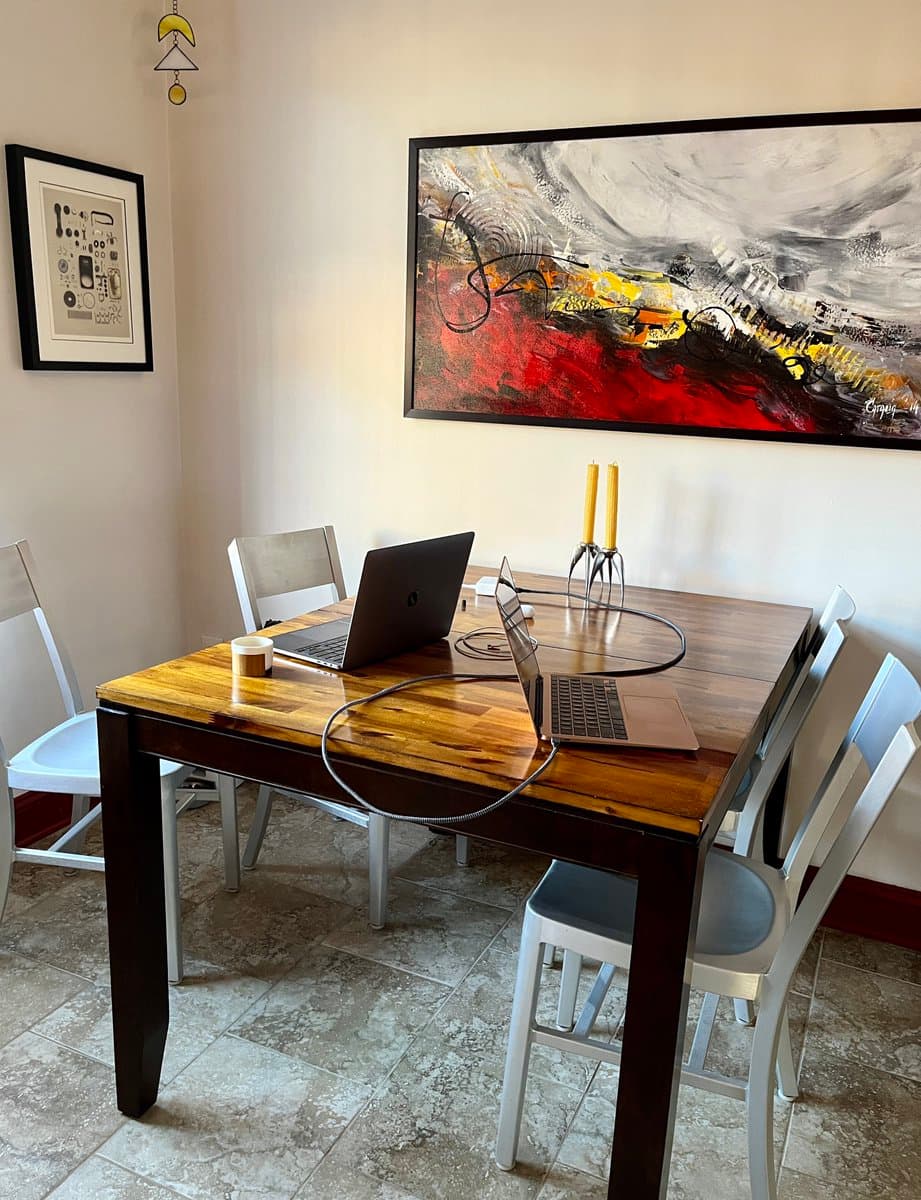 A photo of a dining room table set up with two open laptops, a coffee cup, and a coiled cable, with two yellow candles in a holder on the table. A large abstract painting hangs on the wall behind the table, and a framed print is on the left wall.