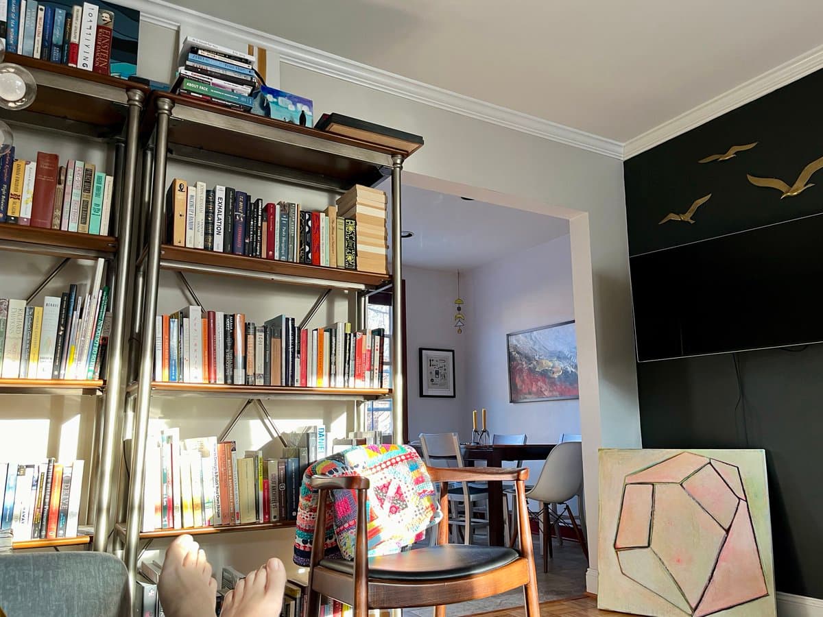 A first-person perspective photo of a person relaxing in a living room with their bare feet propped up, looking towards a large, full bookshelf and a dining area in the background. The room features a dark accent wall with a mounted television and decorative bird sculptures, along with a colorful quilted blanket on a wooden chair.