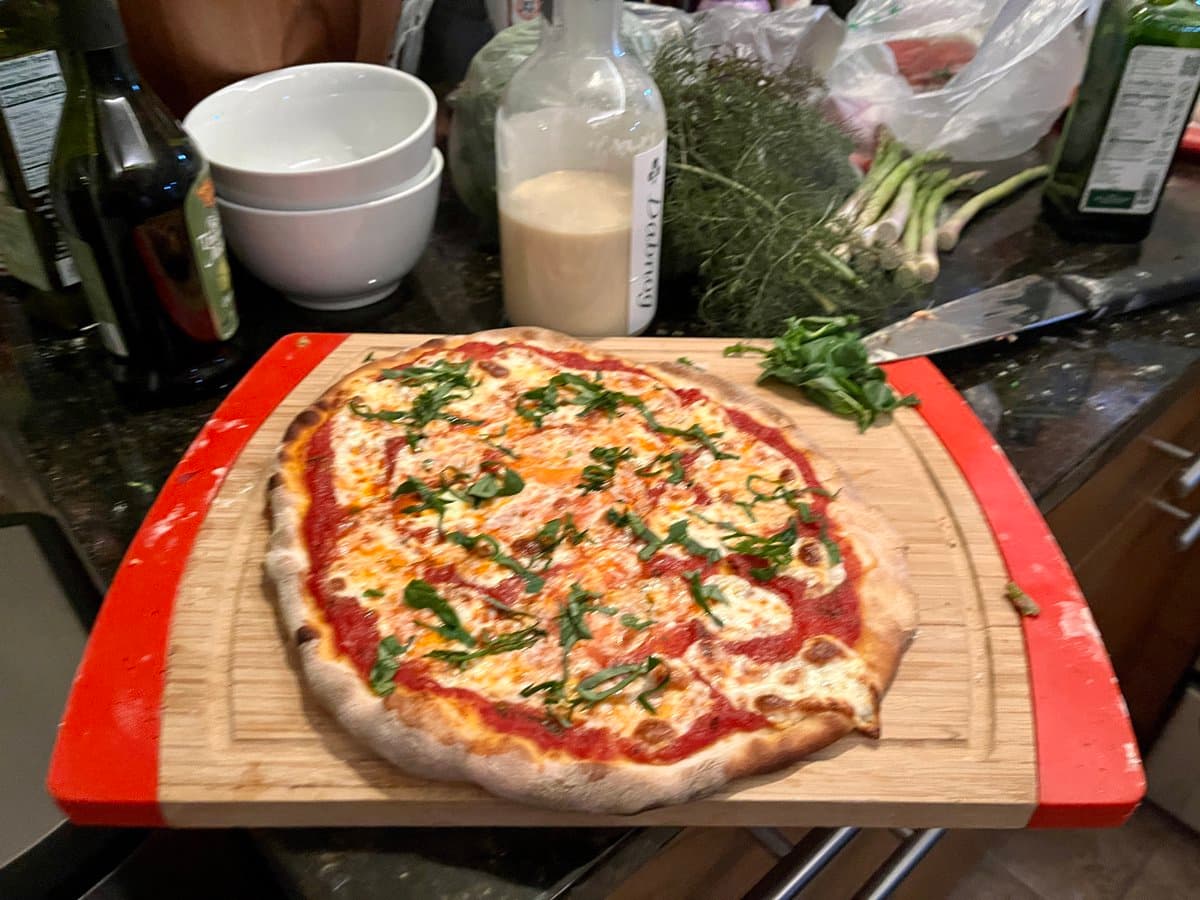 A freshly baked pizza topped with tomato sauce, melted cheese, and fresh basil leaves sits on a wooden cutting board with red edges. In the background, a kitchen counter holds white bowls, a bottle of white wine, fresh herbs, and a knife.