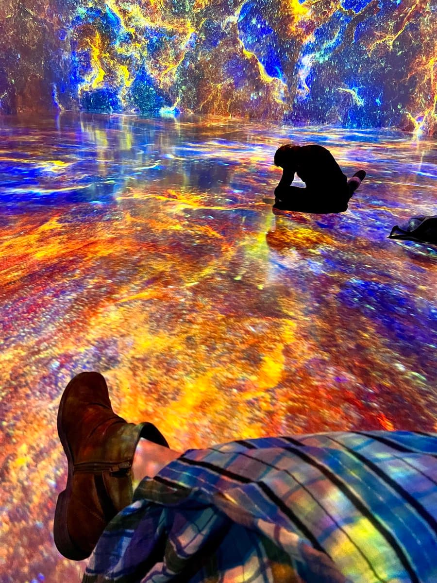 A first-person perspective photo taken inside an immersive digital art installation, showing a person sitting on a reflective floor illuminated by vibrant, swirling projections of blue, yellow, and orange light. The foreground features the photographer's legs and brown shoes, while another visitor is visible in the distance, silhouetted against the glowing walls.