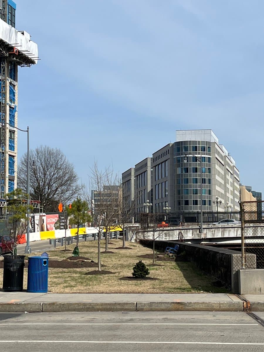 A street-level view of a modern office building with a curved glass corner, situated across a road and a small grassy area with young trees. To the left, a taller building is under construction, covered in blue protective sheeting and scaffolding.