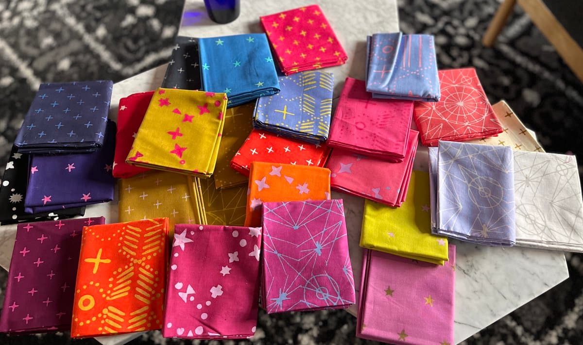 A collection of colorful, folded fabric squares with various geometric and celestial patterns is arranged on a white marble table. The fabrics feature designs such as stars, crosses, and abstract lines in a vibrant palette of pinks, blues, yellows, and oranges.