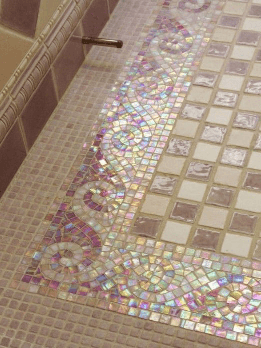 A close-up photograph of a tiled floor featuring a decorative border made of small, iridescent mosaic tiles arranged in a spiral pattern. The mosaic border is set against a background of larger, square tiles in shades of beige and brown.
